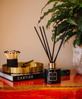 Load image into Gallery viewer, Aromatherapy diffuser with black sticks on a wooden surface with a book and plant in the background.