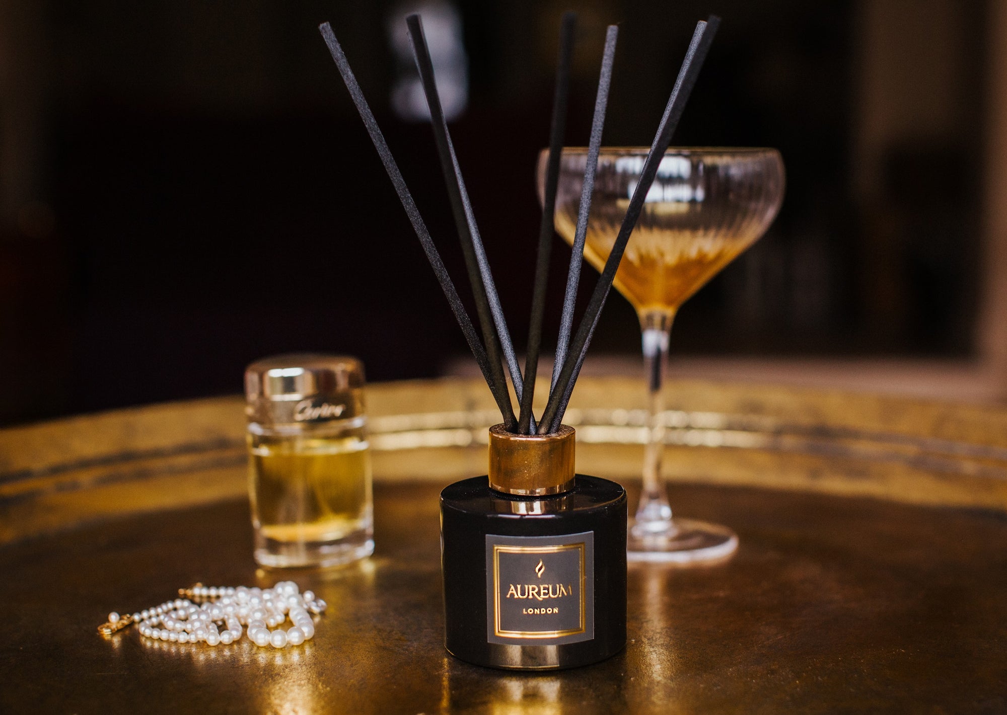 Aurelia diffuser with black reeds on a wooden surface with a glass of cocktail and jewelry.