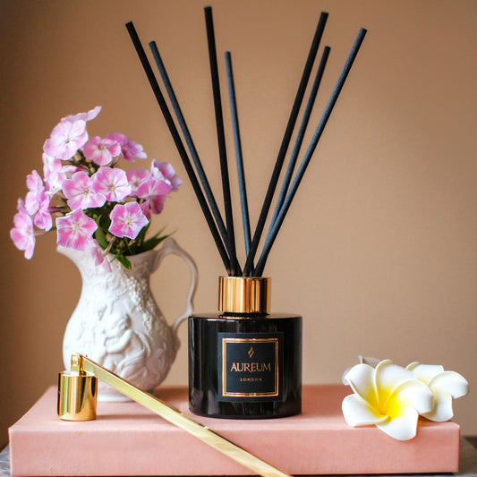 Aromatherapy diffuser with black sticks on a pink stand, surrounded by flowers and a candle.